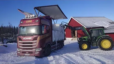 Traktor laster rundballer inn i rød lastebil utenfor låve i snø.