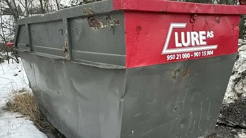 Grå avfallscontainer med rød kant og logoen Lure AS i snødekt terreng.