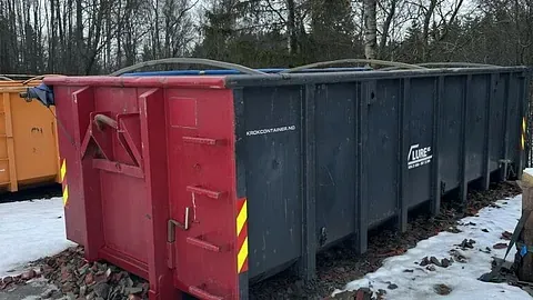 Stor mørk avfallscontainer med rød ende, parkert ute på snø.