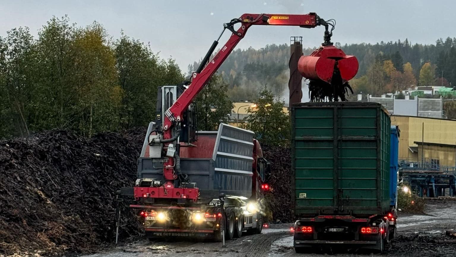 Lastebil med kran som fyller opp en annen lastebil med avfall