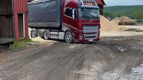 Rød lastebil med sort tippkasse rygger ved sagbruk og flishaug.